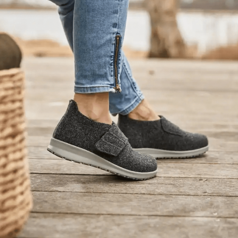 Donna che indossa pantofole New Feet grigio antracite su un pavimento in legno, vista laterale dei piedi