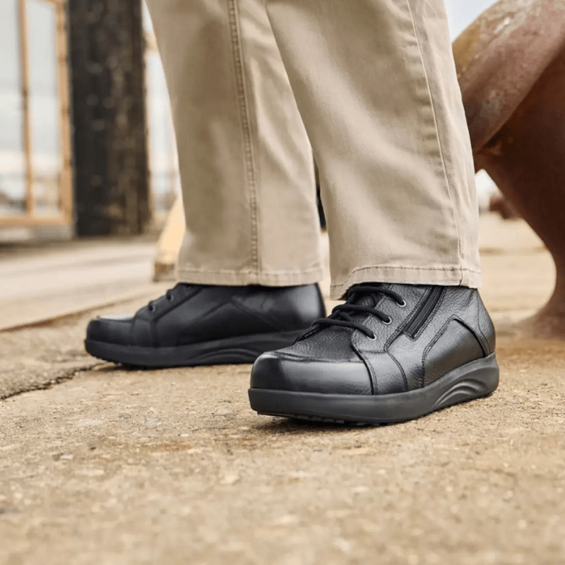 Un uomo con pantaloni beige e stivaletti New Feet neri da uomo in piedi su una superficie grigia