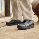 Un uomo con pantaloni beige e stivaletti New Feet neri da uomo in piedi su una superficie grigia